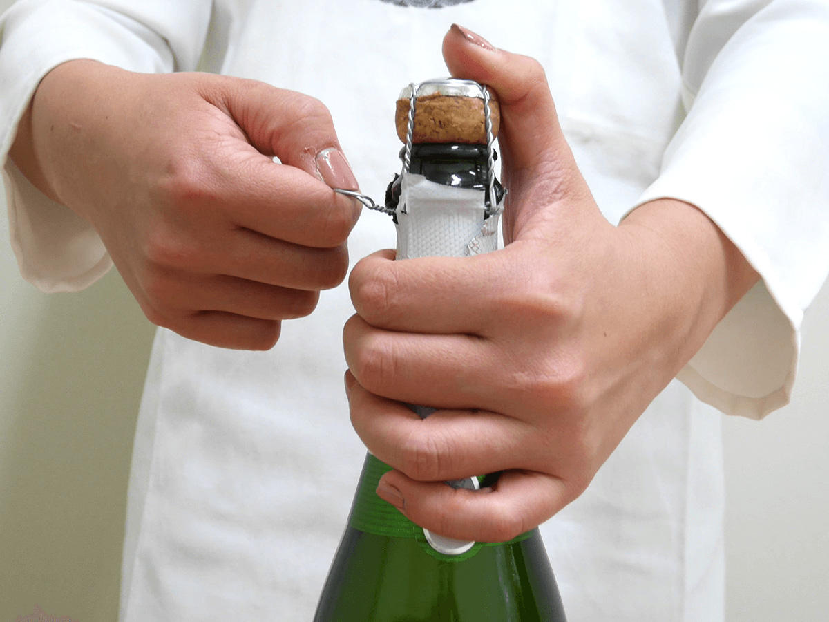シャンパン、スパークリングワインの開け方解説の写真２。片手で瓶の首を握り、親指で栓をしっかり押さえながら、針金をゆるめる。
